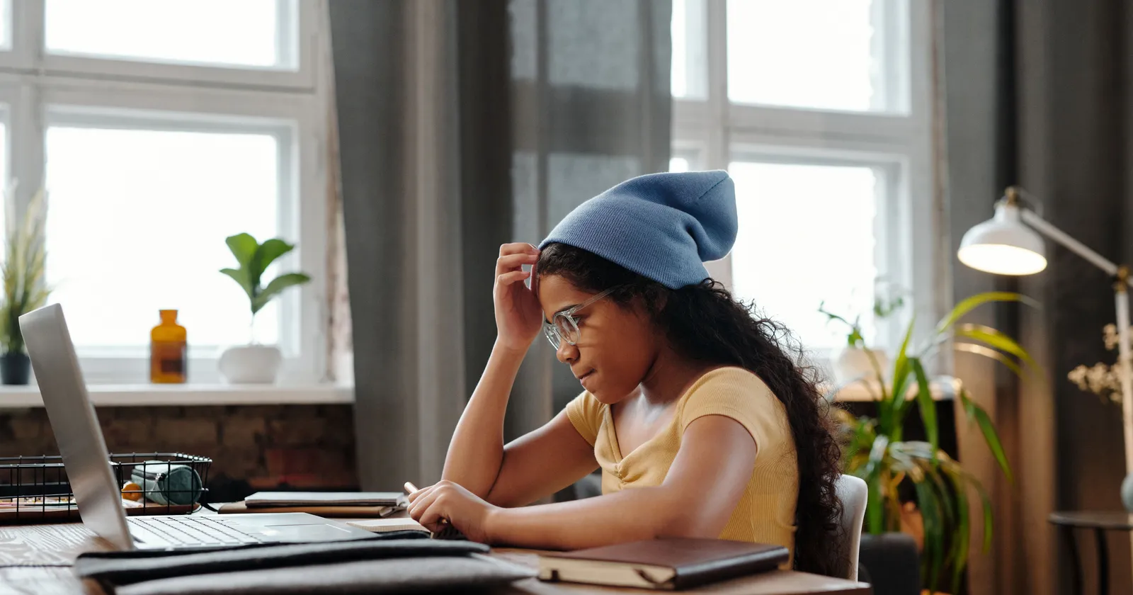A girl in glasses and a blue beanie studying at a laptop at a desk by a window