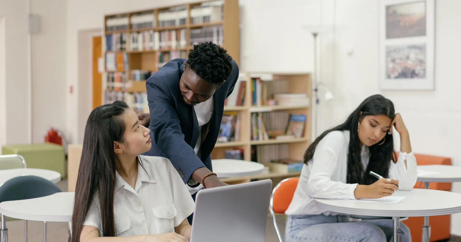 A teacher leaning over a student's laptop to help during class, with another student writing at the next table