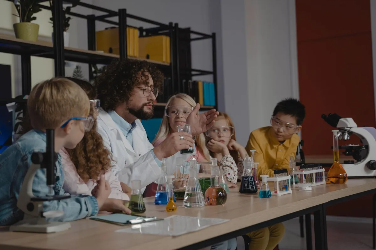 A teacher in a lab coat running a chemistry demonstration with a group of students in safety glasses around a table of flasks